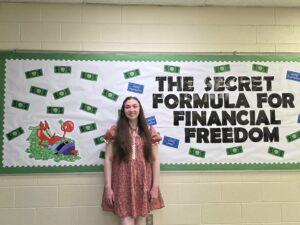 female teacher wearing a red dress standing in front of a bulletin board decorated with paper money, Mr. Krabs sitting on a pile of money, and text that says, "The secret formula for financial freedom."