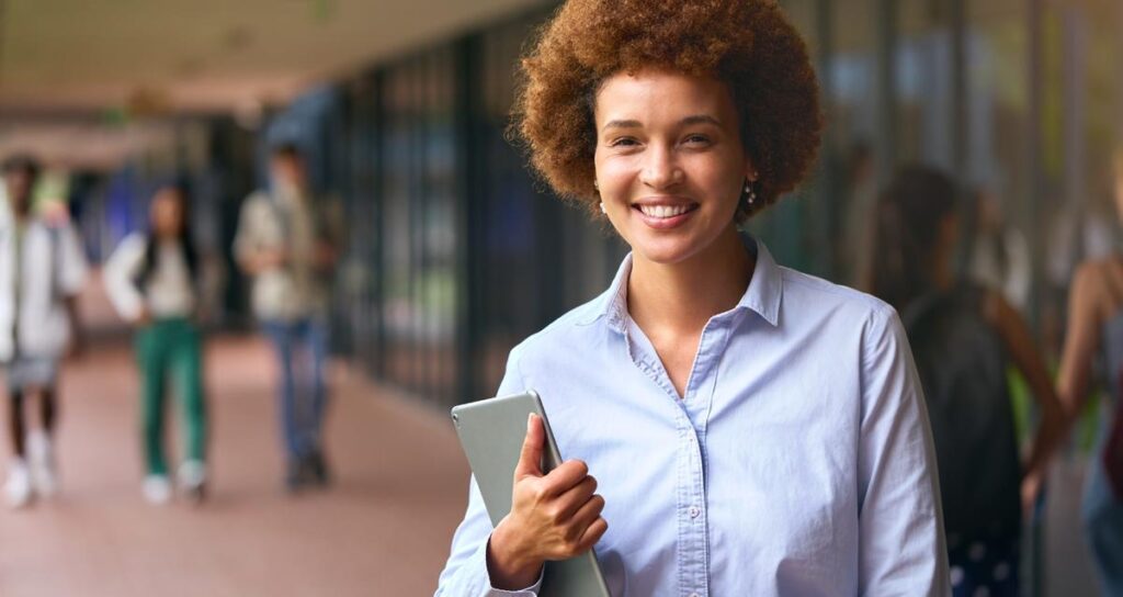 Educator smiling while holding a tablet in a school hallway, representing K12 Coalition’s commitment to teaching for the greater good