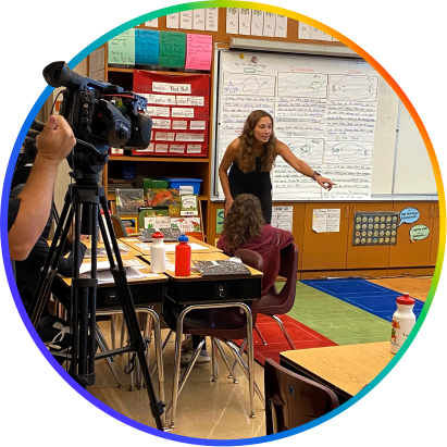 A teacher in a classroom, calling on a student, while a camera records the lesson in the corner.