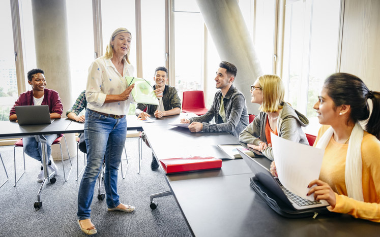 Group of young people listening to female lecturer in FE college, woman with laptop smiling and looking at teacher