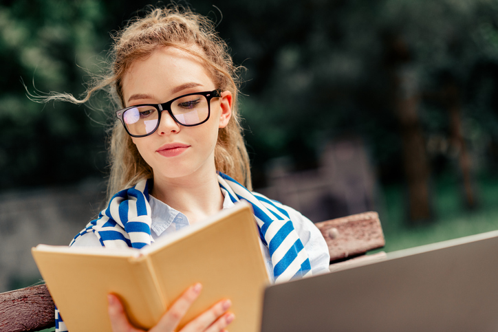 A young woman enjoys reading a book while sitting outdoors in an urban park. She wears stylish eyeglasses and a scarf, reflecting leisure and relaxation on a sunny day.