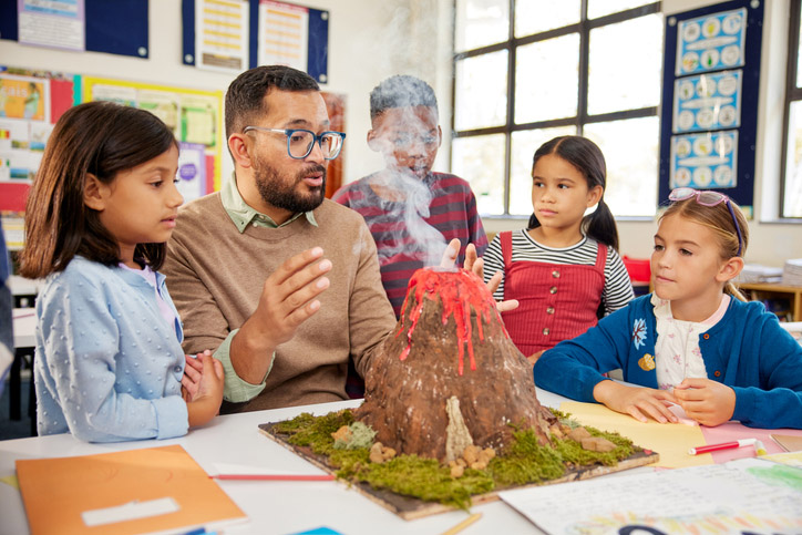 Male teacher teaching to a group of school children about volcanos at science project in school. Group of students understanding volcanic eruption in class with teacher during science class while observing a volcano model that actually erupts with the presence of smoke.