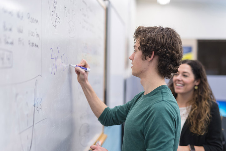 A middle-aged teacher stands at the whiteboard and works with a male high school student on a math problem.